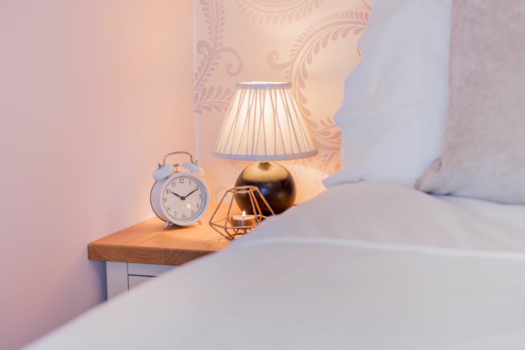 Nightstand in the corner with lamp, clock and a candle on it. 