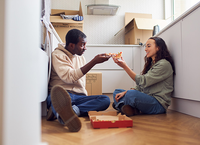 A couple sat on a kitchen floor eating a pizza 