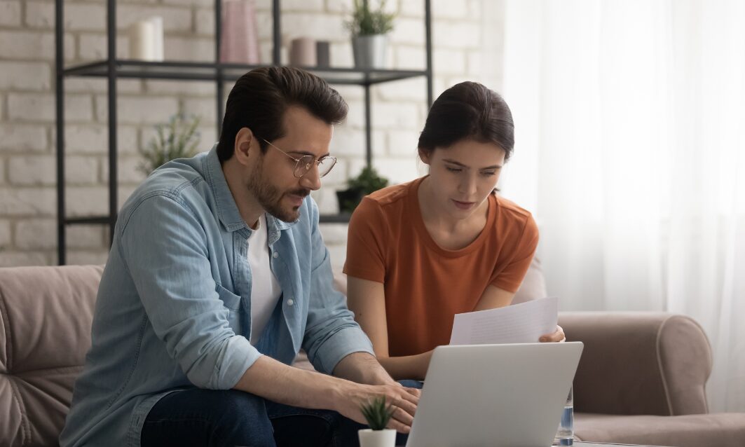 Couple sitting on a sofa reviewing property documents and checking compliance information on a laptop.