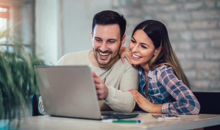Couple smiling together while looking at a laptop, researching homes for sale online at a kitchen table