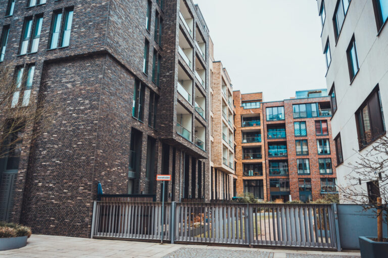 Modern multi-story apartment buildings with dark and red brick facades, large windows, glass balconies, and a metal security gate enclosing a paved courtyard.