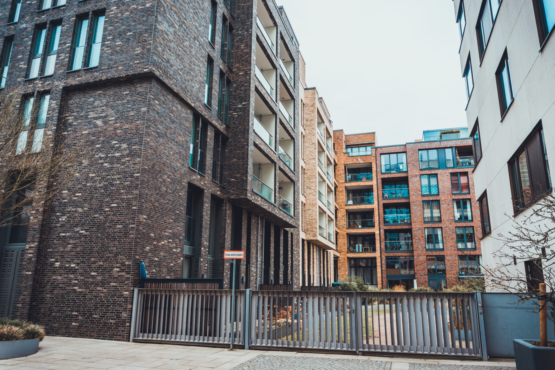 Modern multi-story apartment buildings with dark and red brick facades, large windows, glass balconies, and a metal security gate enclosing a paved courtyard.