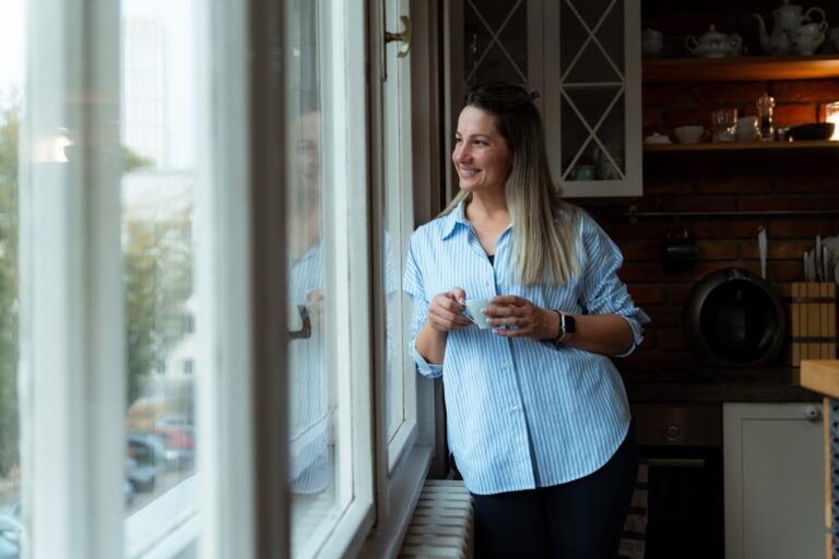 Woman holding a cup of coffee and looking out of a kitchen window, representing village life in Dilton Marsh BA13 near Westbury