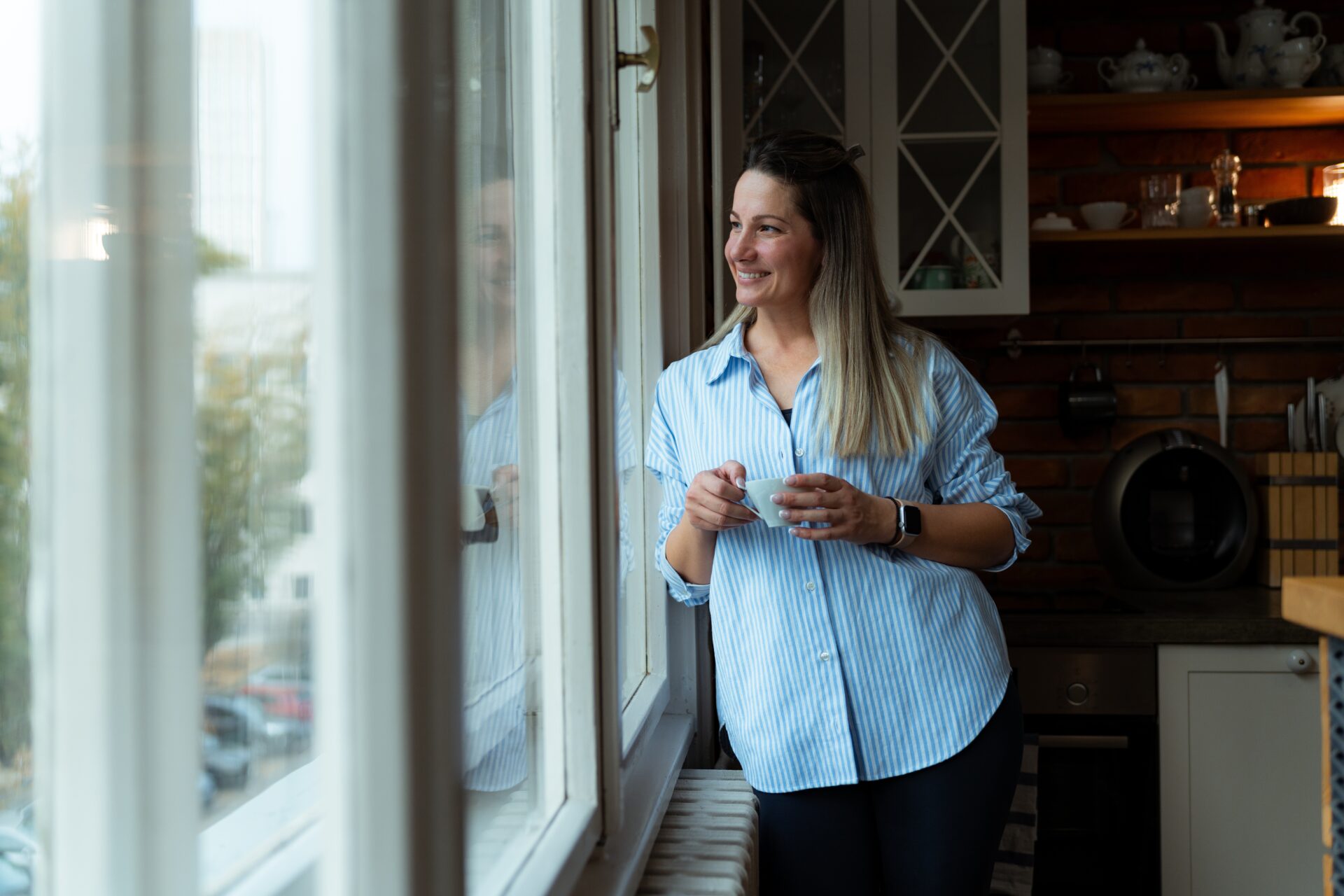 Woman holding a cup of coffee and looking out of a kitchen window, representing village life in Dilton Marsh BA13 near Westbury