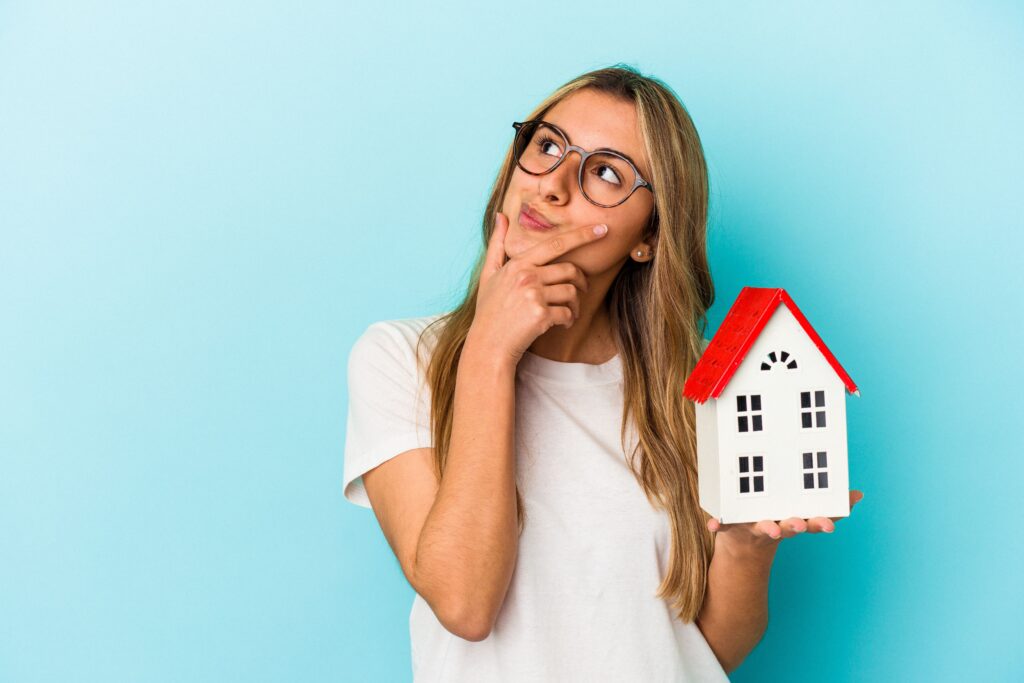 Young caucasian woman holding a house model isolated on blue background looking sideways with doubtful and skeptical expression.